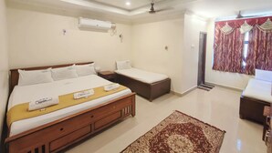 A hotel room with a large bed, a smaller bed, a desk, and a wardrobe. There is a ceiling fan, an air conditioning unit, and a patterned rug on the floor. - Hotel Harish (Rameshwaram)