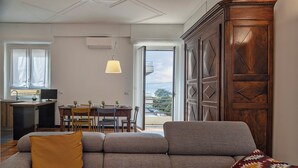 Ein modernes Wohnzimmer mit einem grauen Sofa, einem gelben Kissen und einer gemusterten Decke. Im Hintergrund ist eine Essecke mit einem Holztisch und Stühlen zu sehen.