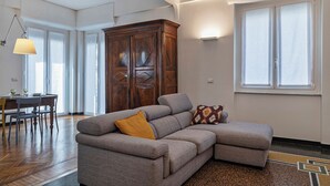 Ein Wohnzimmer mit einem grauen Ecksofa, einem Holzschrank, einem Esstisch mit Stühlen und einem großen Fenster.