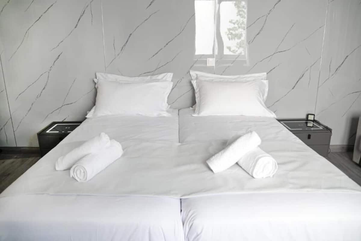Un lit double avec du linge de lit blanc et deux serviettes enroulées, une table de chevet avec un téléphone, et un mur décoré d’un motif imitant le marbre.