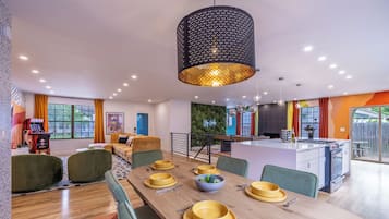 A modern dining area with a long table set for four, a kitchen with a gas stove, and a living area with a sofa and a gaming console.