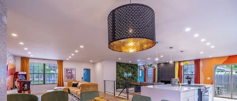 A modern dining area with a long table set for four, a kitchen with a gas stove, and a living area with a sofa and a gaming console.
