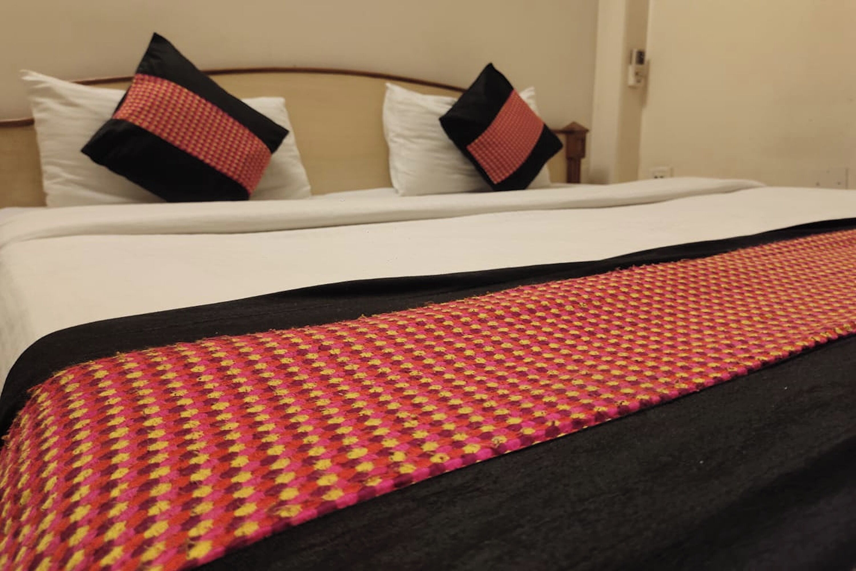 A hotel room with a bed, white bedding, and a red and black patterned throw blanket.