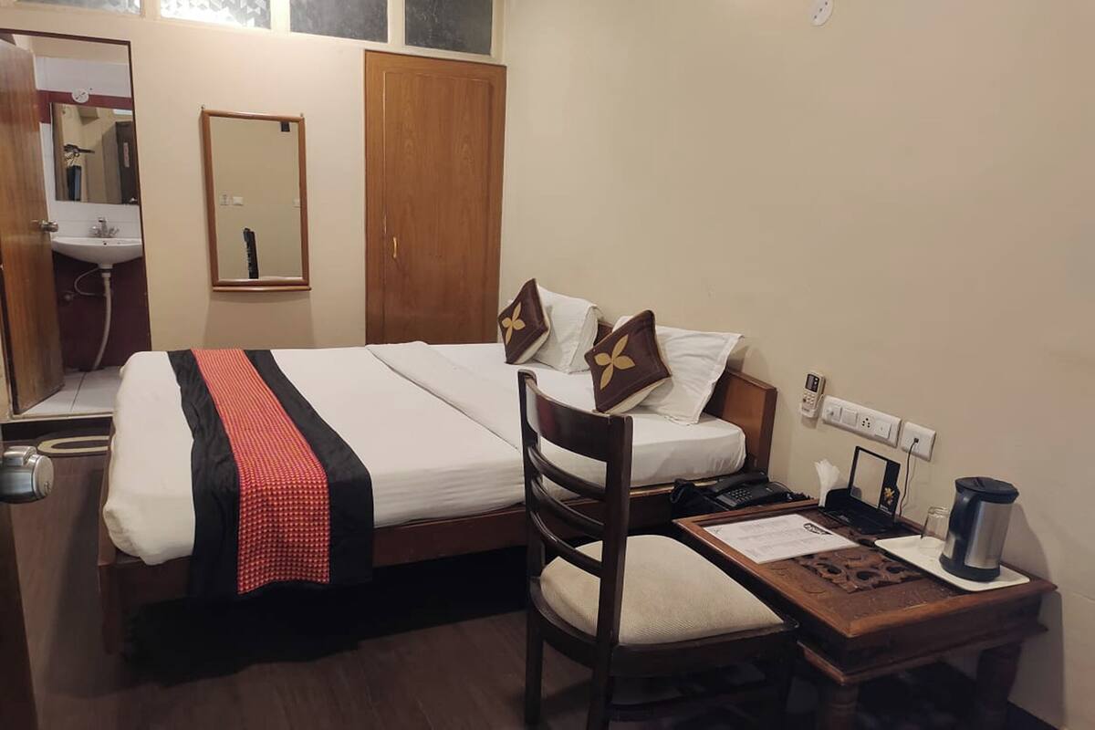 A hotel room with a bed, a desk with a laptop, a chair, a mirror, a wardrobe, and a sink.
