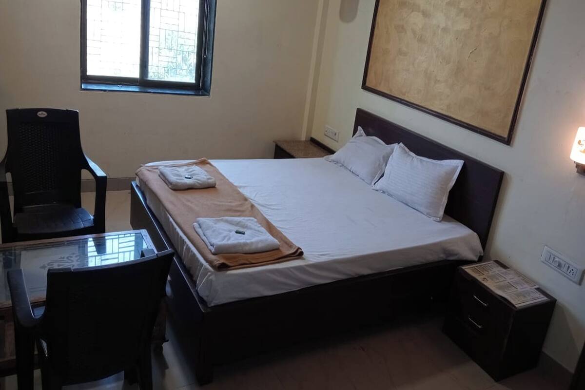 A hotel room with a single bed, a small table with a chair, a window, and a wall-mounted light.