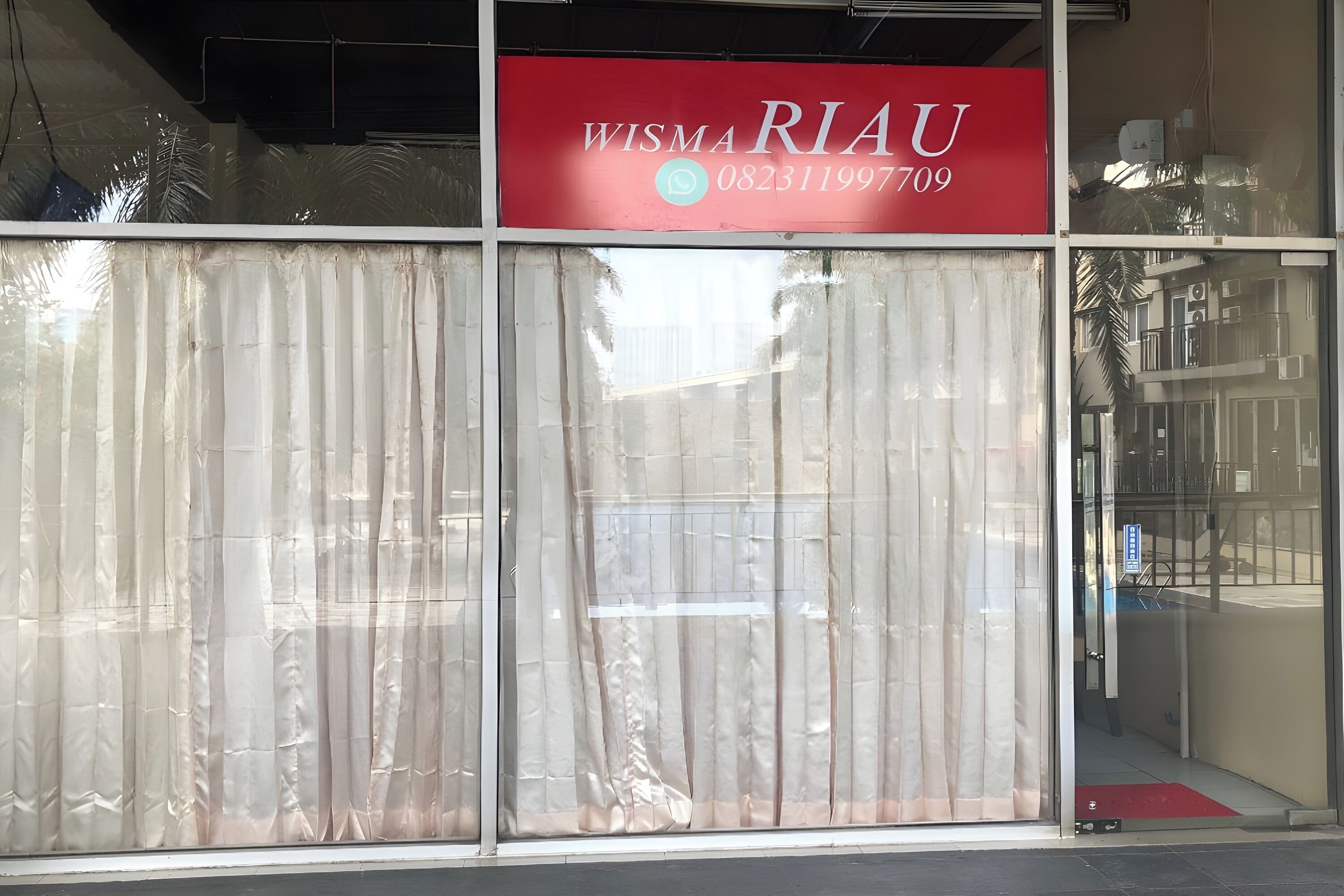 Fachada de un comercio con un letrero rojo que dice "WISMA RIAU" y un número de contacto.