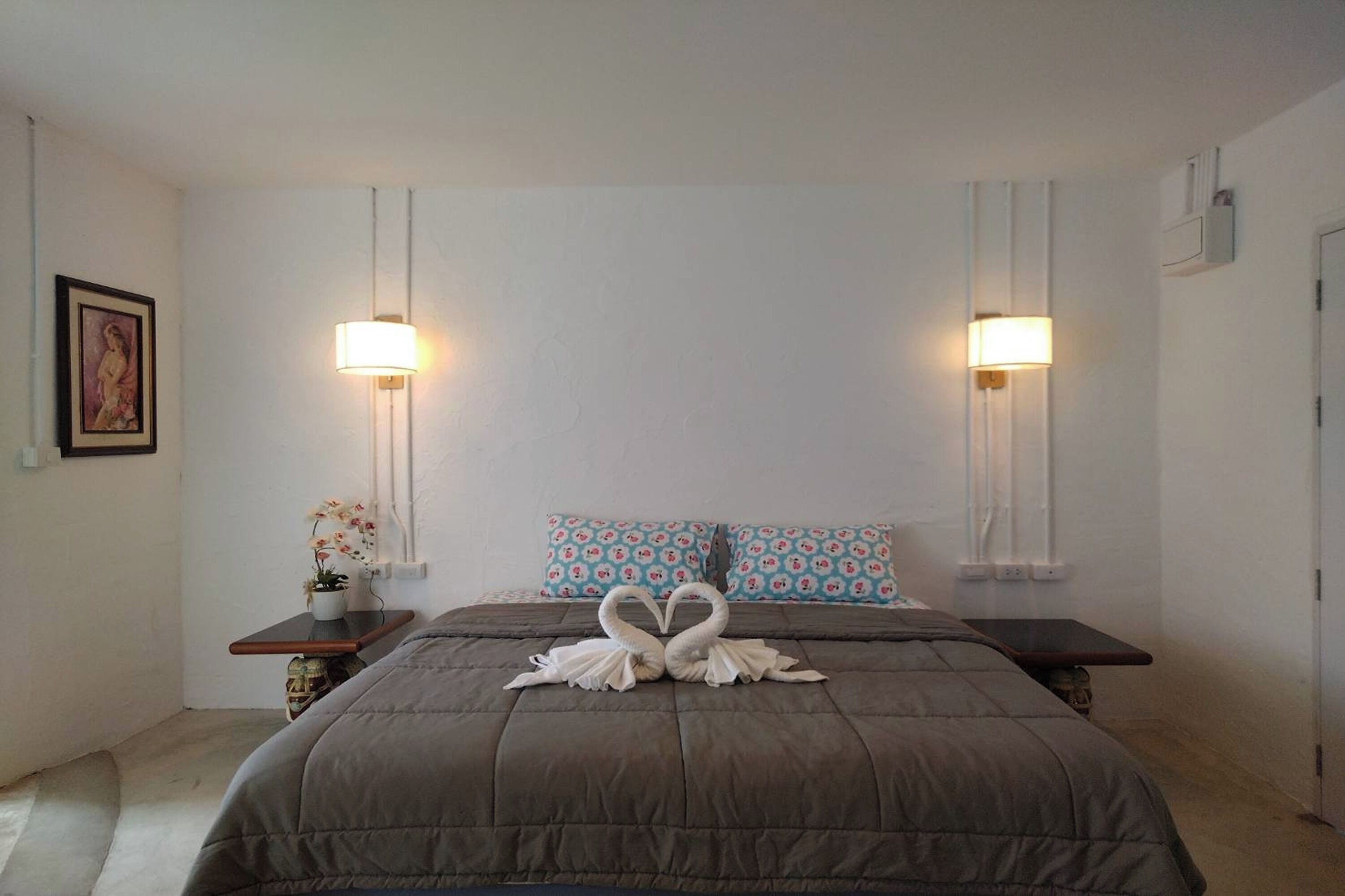 A bedroom with a large bed, two bedside tables, a wall-mounted lamp, a framed picture, and patterned pillows.