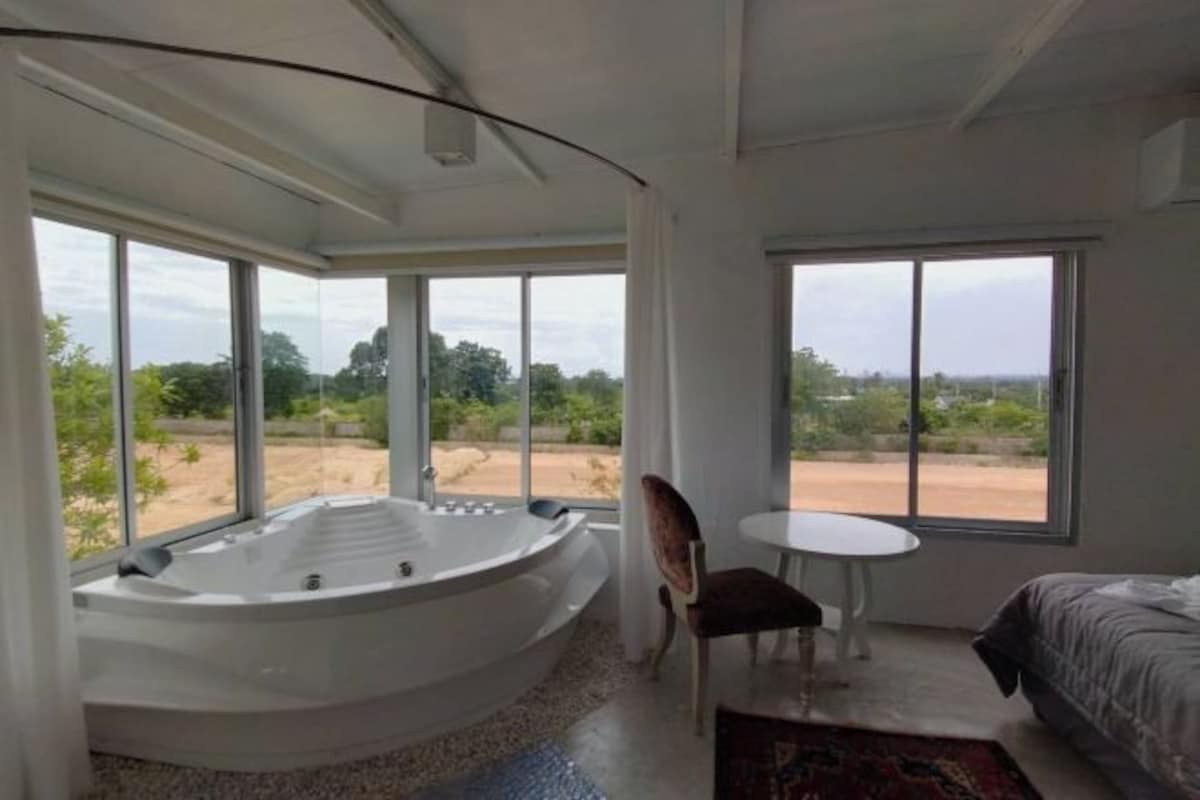 A room with a large bathtub, a bed, a small table, and a chair. There are large windows providing a view of the outdoors.