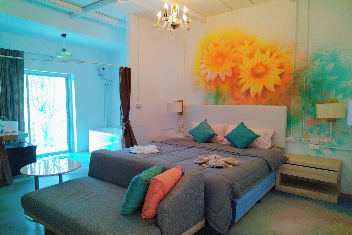 Quarto com cama grande, sofá, mesa redonda e parede com mural de girassol.