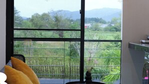 Una sala de estar moderna con un sofá, una mesita y un amplio ventanal corredera que da a un balcón con vistas a zonas verdes y montañas.