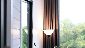 Una habitación con una puerta blanca, una ventana con vistas a árboles, una planta en maceta y una lámpara de pie.