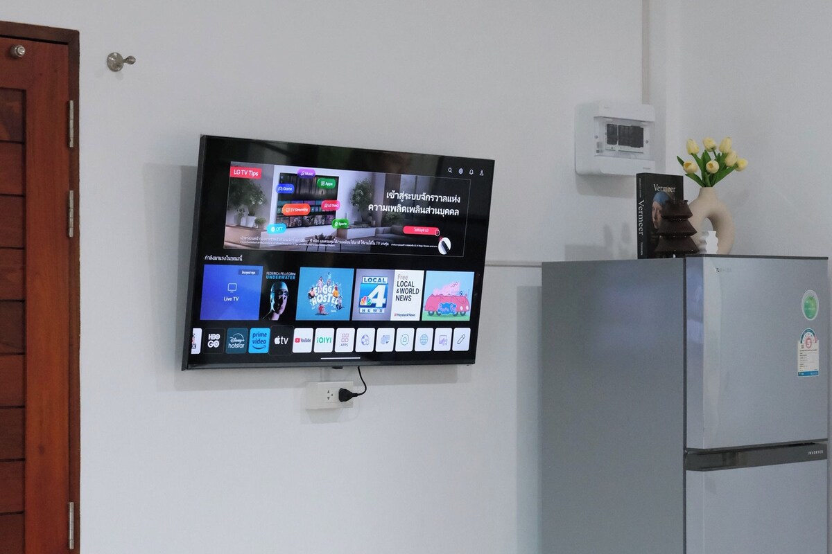 Una habitación con un televisor de pantalla plana montado en la pared que muestra varios canales y un reloj digital. Un refrigerador está colocado contra la pared a la derecha del televisor.