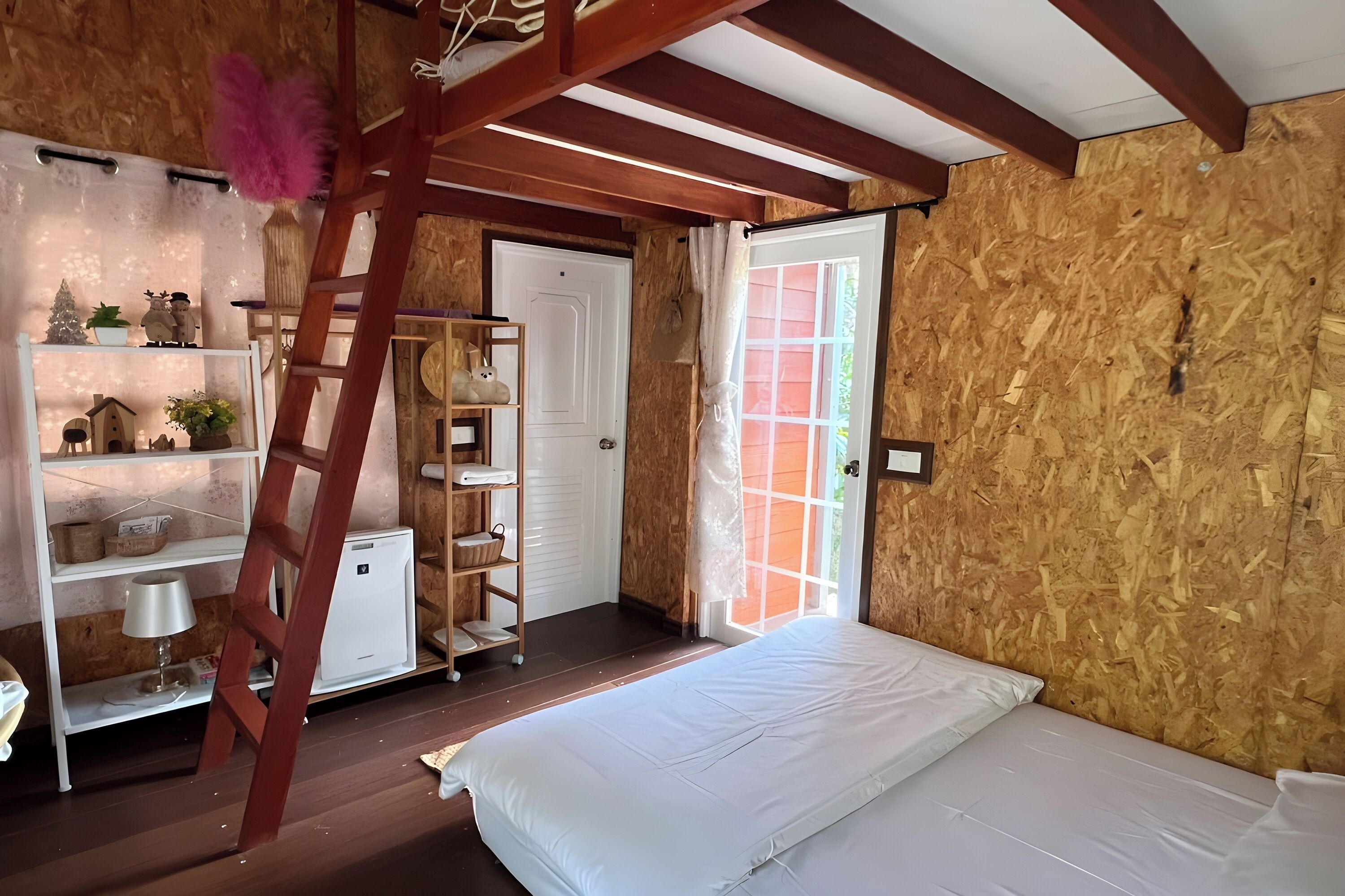 A room with a bed, wooden walls, a ladder, a white door, and a shelf with various items.