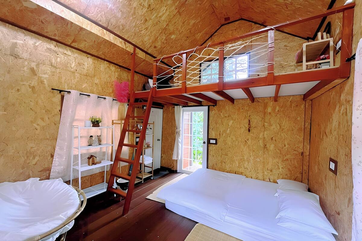 A wooden interior with a bunk bed, a ladder, a white shelf with decorative items, and a door leading outside.