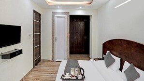A hotel room with a bed, a TV, a ceiling fan, and a door leading to a wardrobe. - Bellmore Stay & Homes (Gurugram)