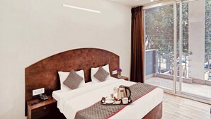 A modern hotel room with a large bed, a bedside table with a phone, a tray with breakfast items, and a view of the outdoors through a sliding glass door. - Bellmore Stay & Homes (Gurugram)