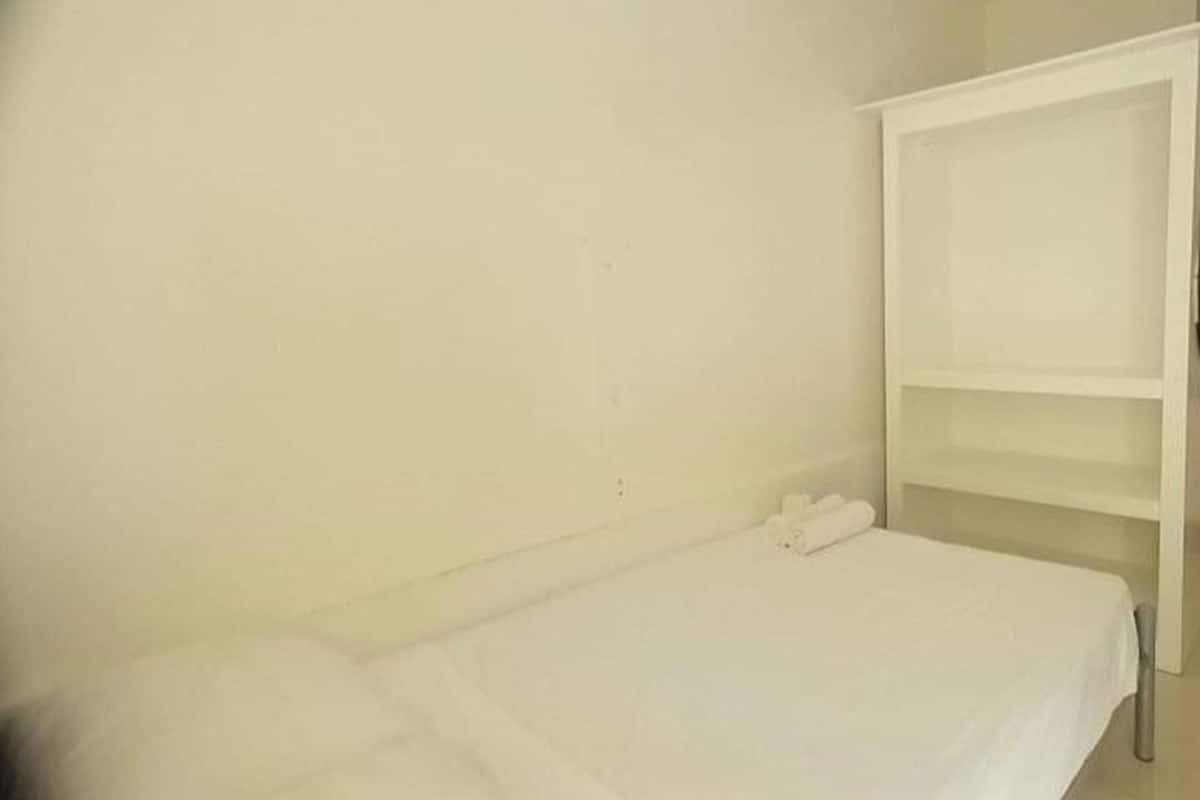 A single bed with a white mattress and a white blanket, a white bedside table, and a white wardrobe with open doors.