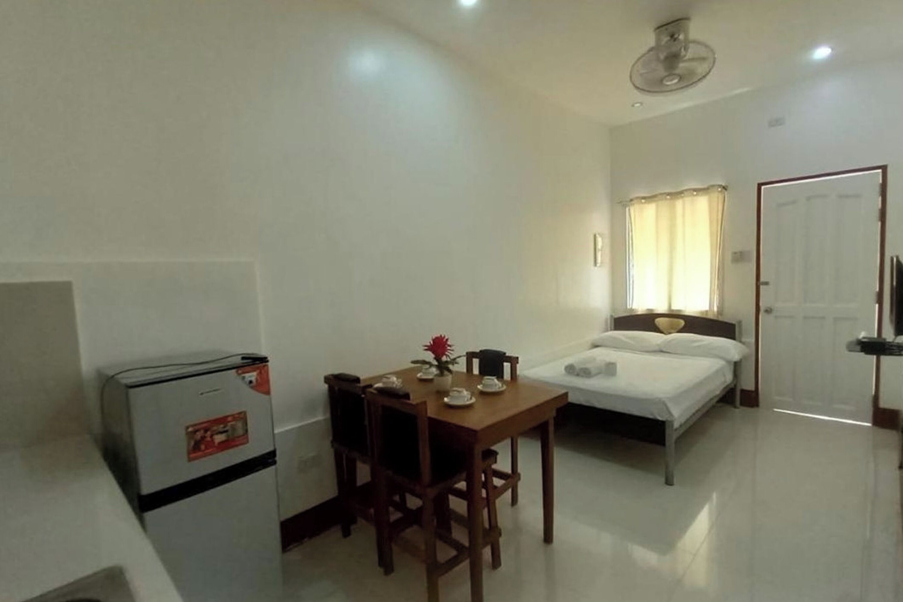 A compact room with a bed, a small table with a flower, a refrigerator, and a ceiling fan.