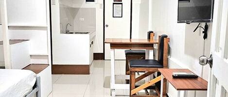 A compact hotel room with a bed, a desk, a chair, a TV, and a small kitchenette.