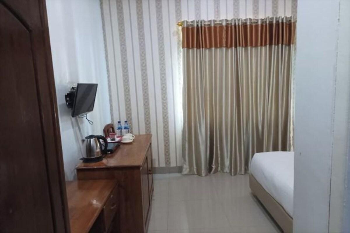 A hotel room with a bed, a desk with a kettle, a TV, and a curtain.