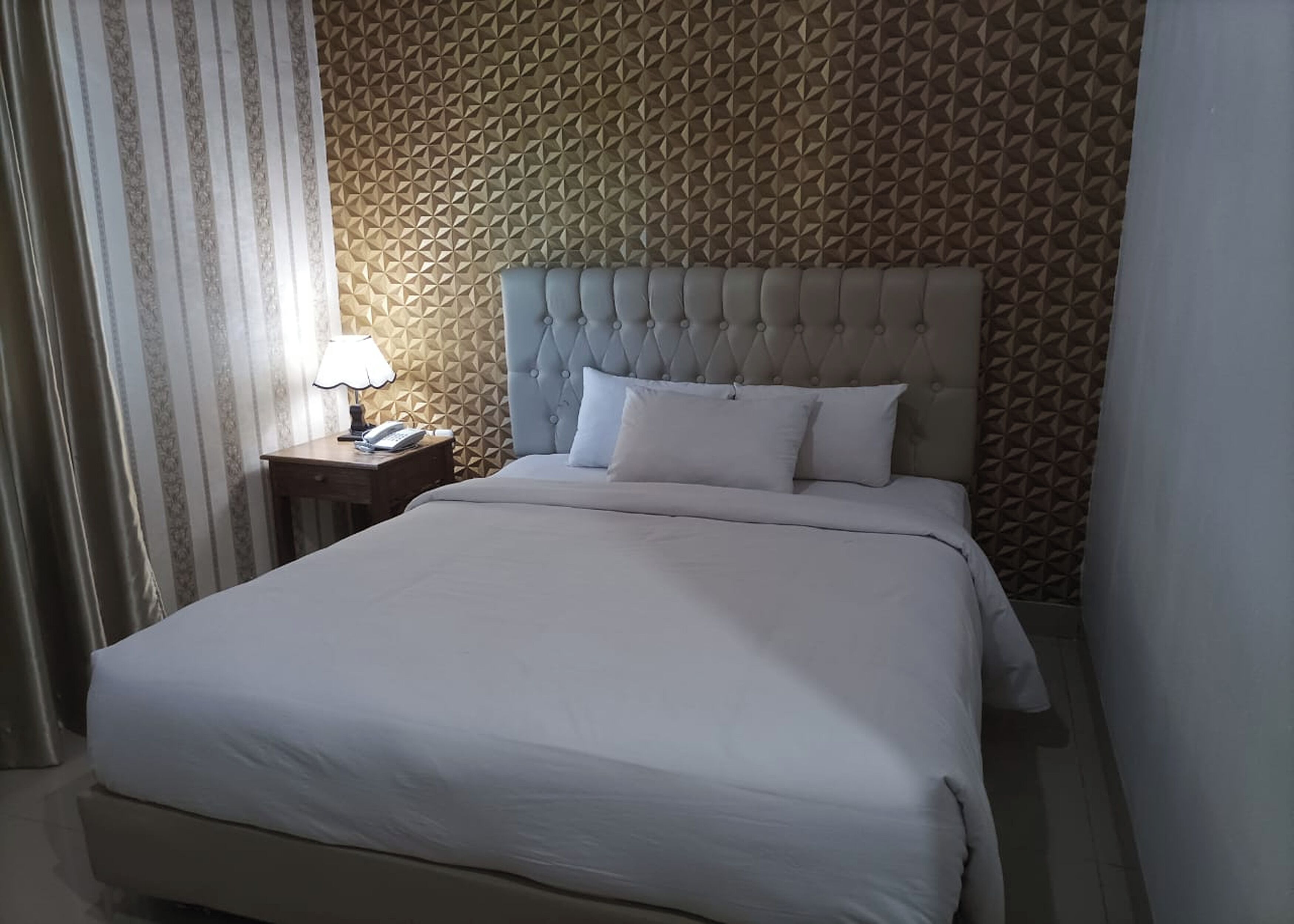 Una cama bien tendida con un cabecero capitonado, una mesita de noche con lámpara y un papel tapiz estampado.