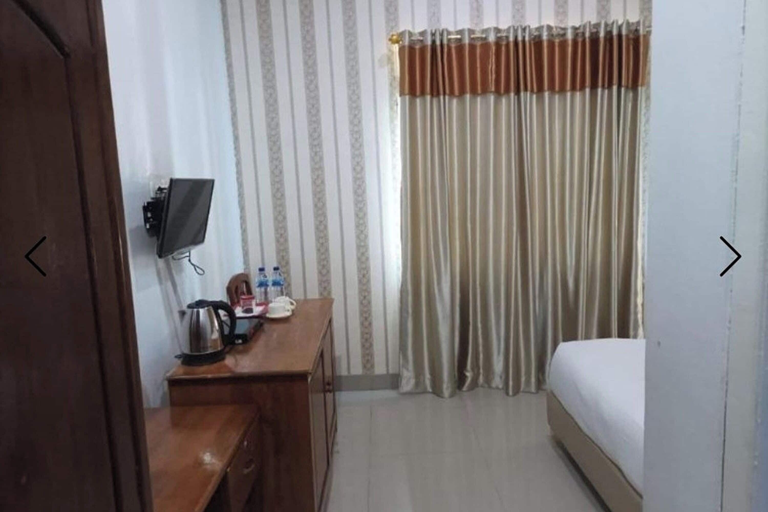A hotel room with a bed, a desk with a kettle, a television, and a window with curtains.
