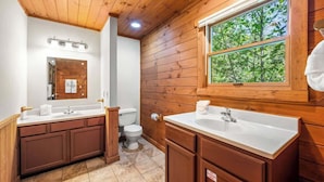 Bagno di una baita in legno con due lavabi, un water e una finestra vista alberi.