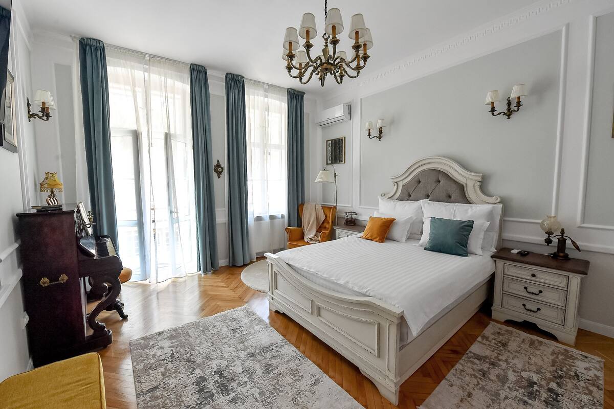 A bedroom with a large bed, a chandelier, a piano, and a dresser.
