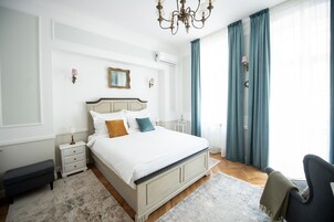 Un dormitorio con una cama grande, mesitas de noche, un banco, una araña y vistas al exterior.