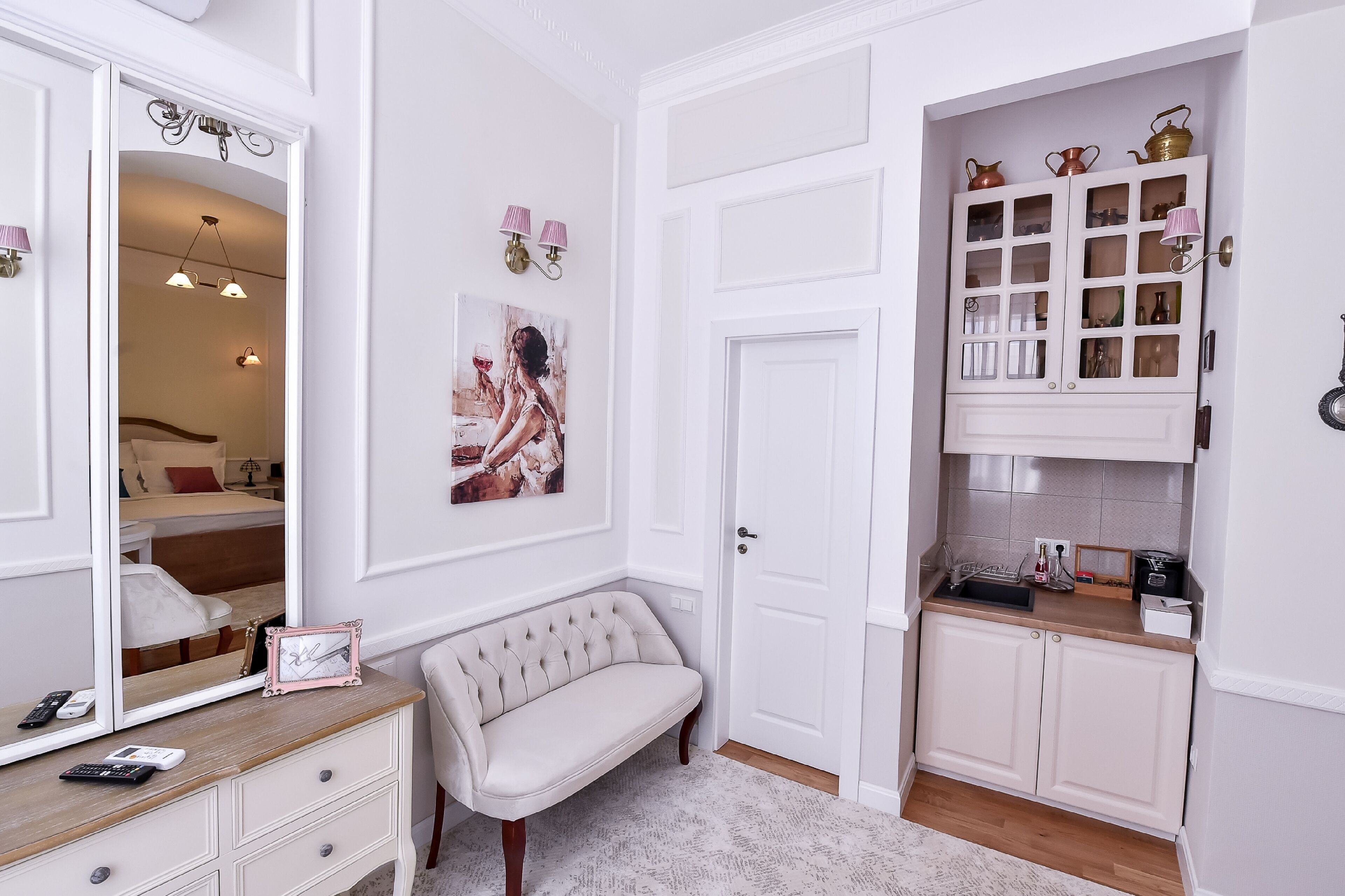 A well-lit bedroom with a large mirror, bedside table, and a bench. There is a wardrobe with a mirror and a cabinet with glass doors.