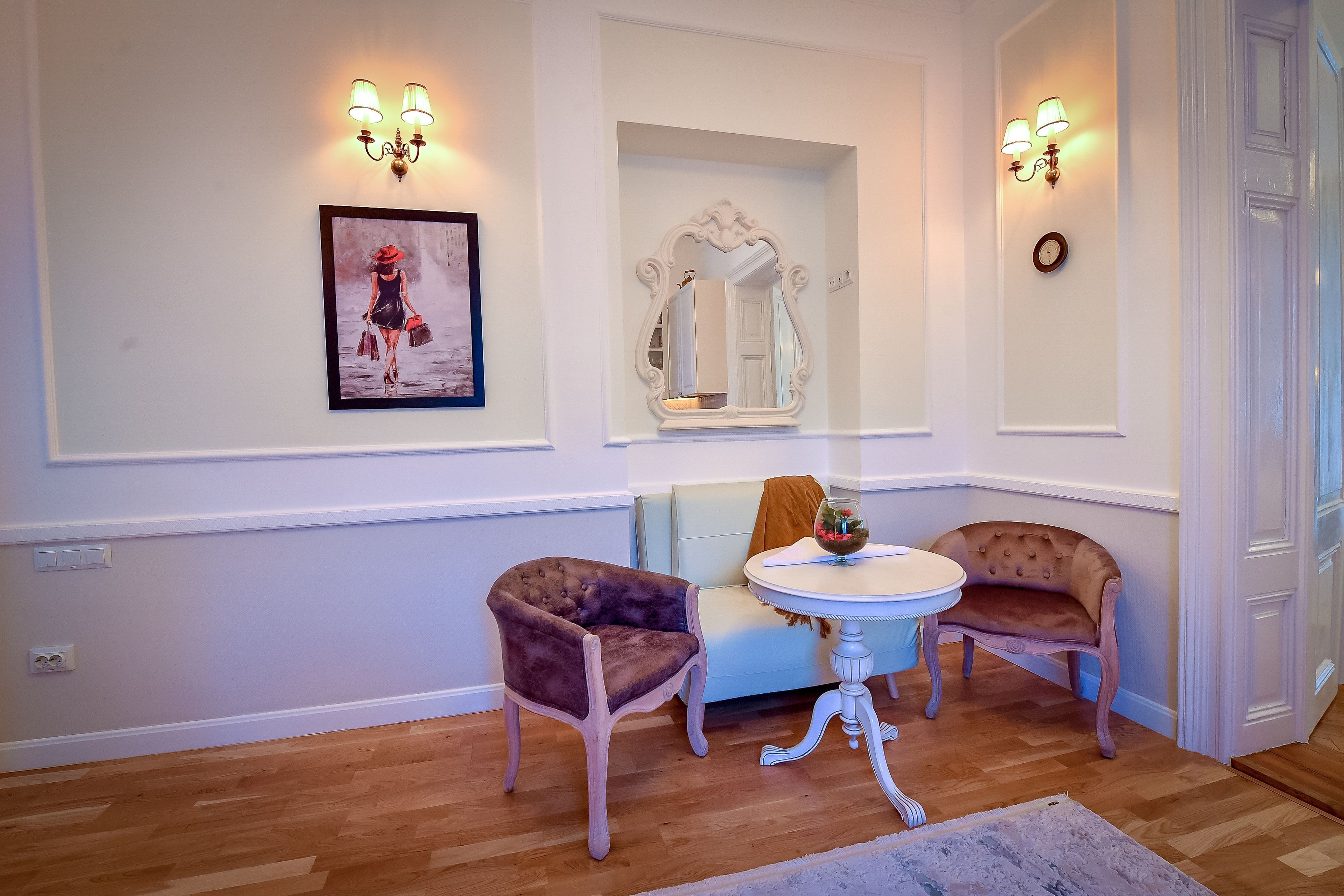 A room with a white wall, a framed picture, a mirror, two upholstered chairs, a small round table, and a light-colored rug.