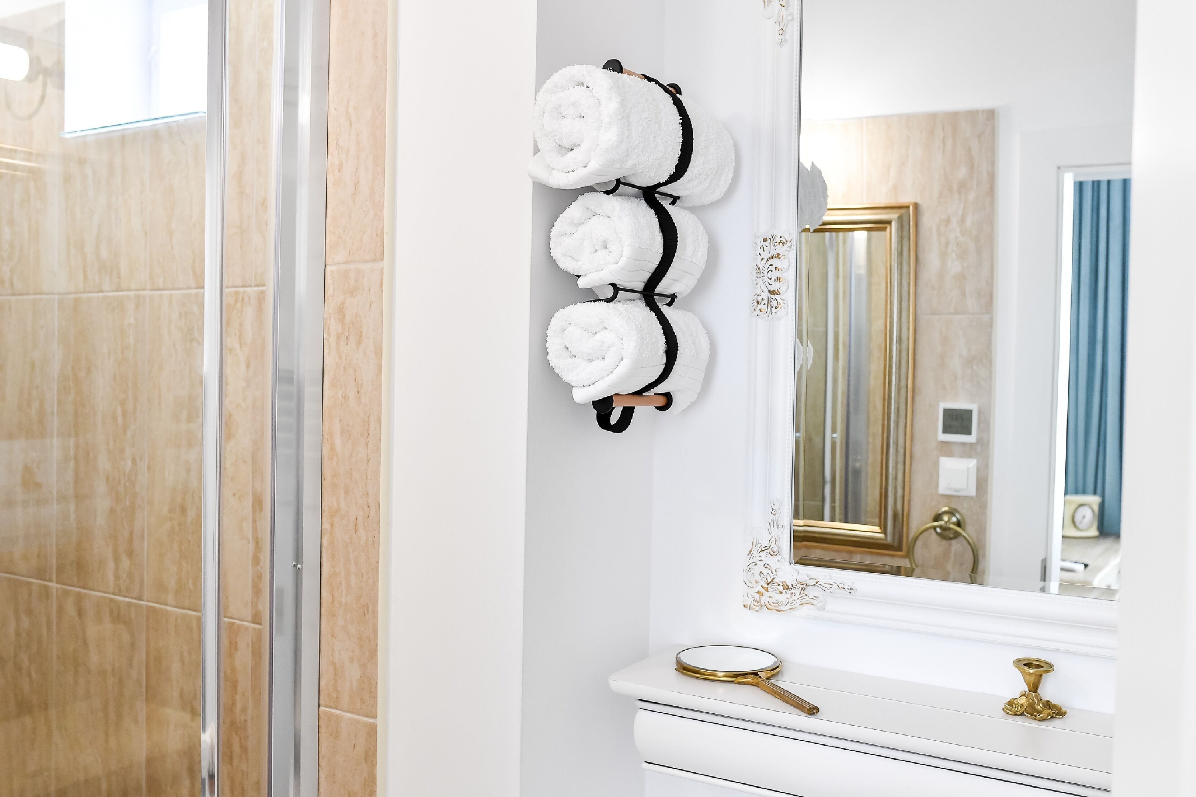 A bathroom with a white vanity, a gold-framed mirror, and three rolled towels on a black towel rack.