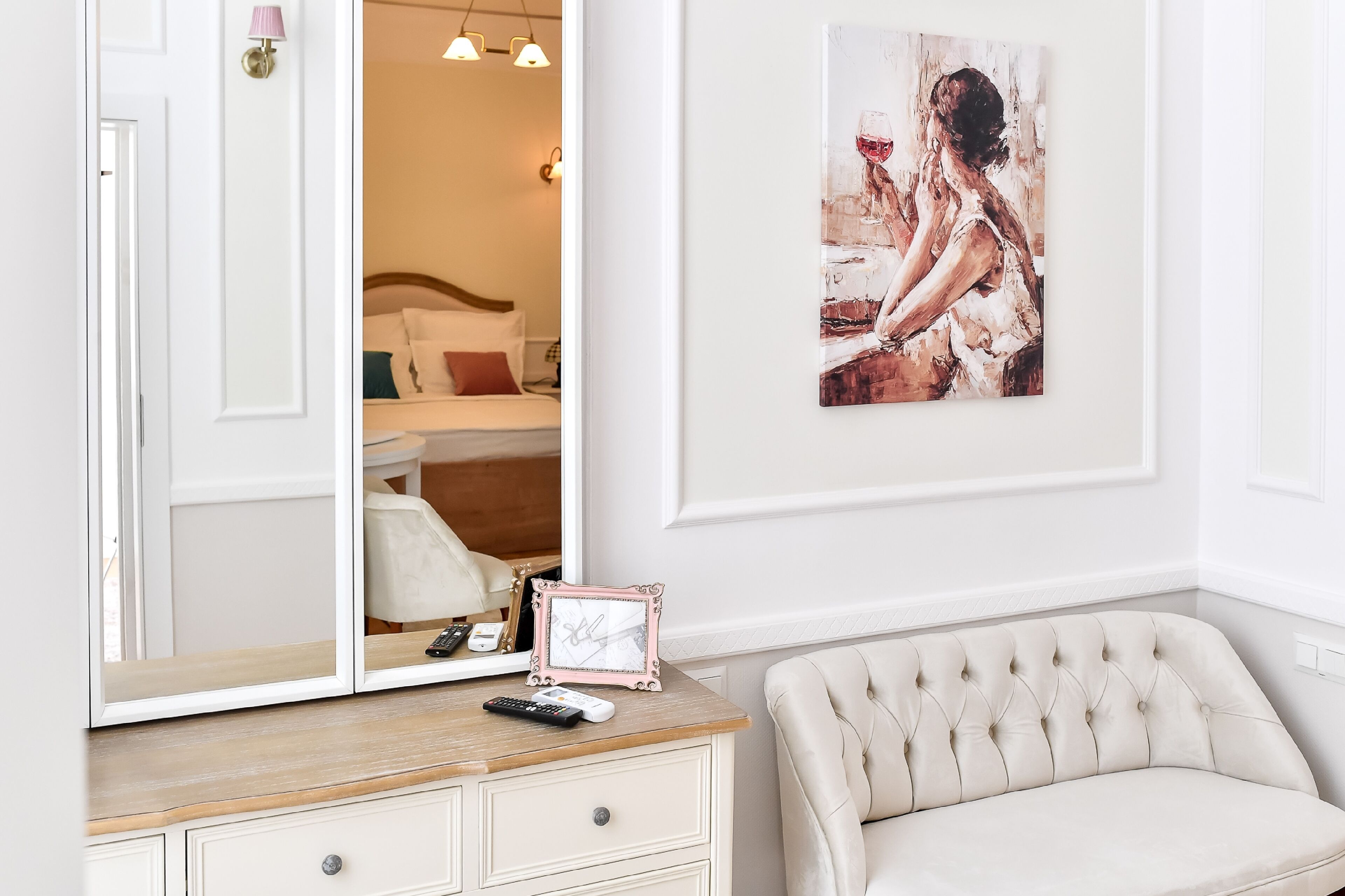 A bedroom with a large mirror, a bedside table with a remote control, a framed picture, and a tufted armchair.