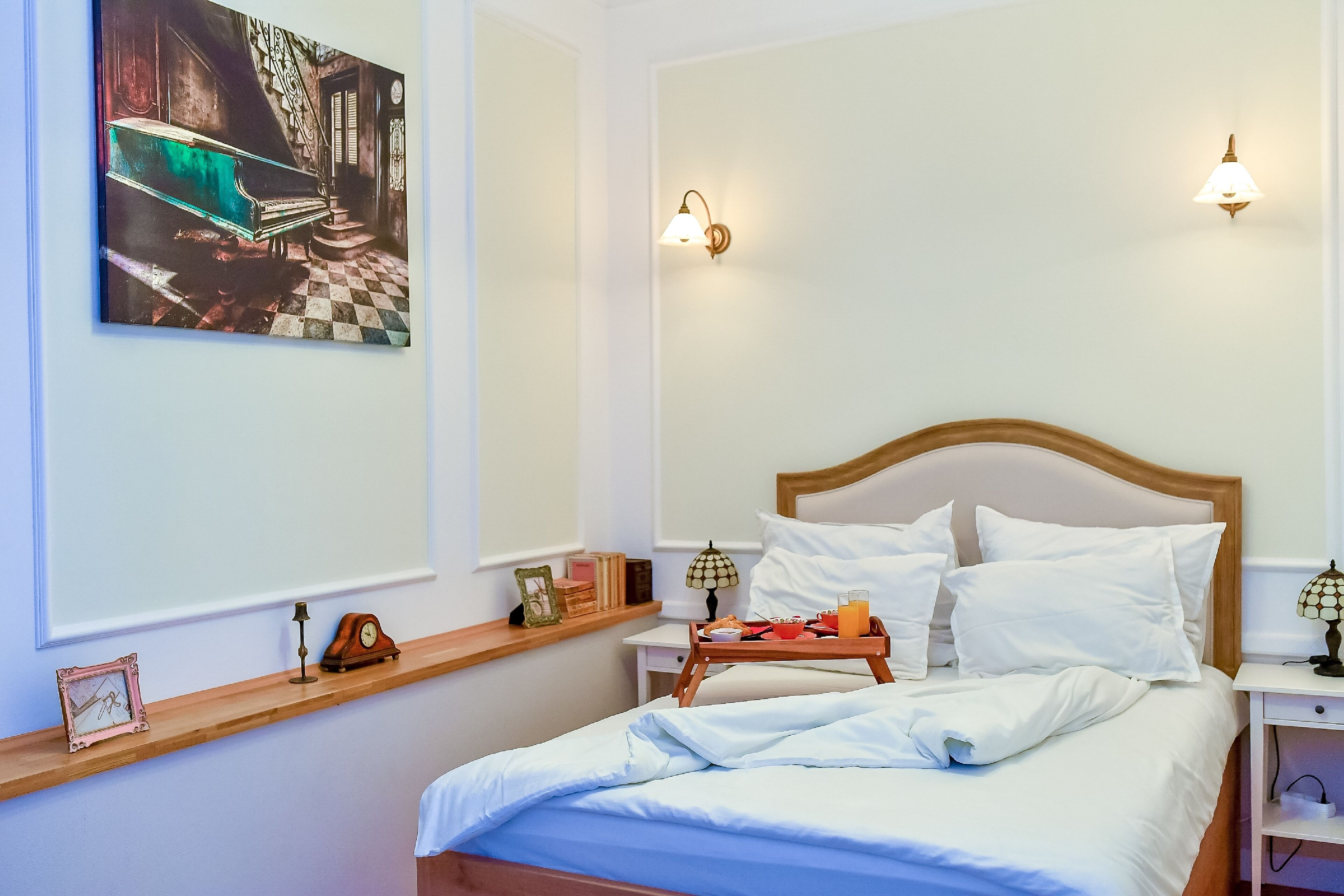 Una cama bien hecha con sábanas blancas, una mesita de noche con lámpara, una bandeja con alimentos para el desayuno y un cuadro sobre la cama.