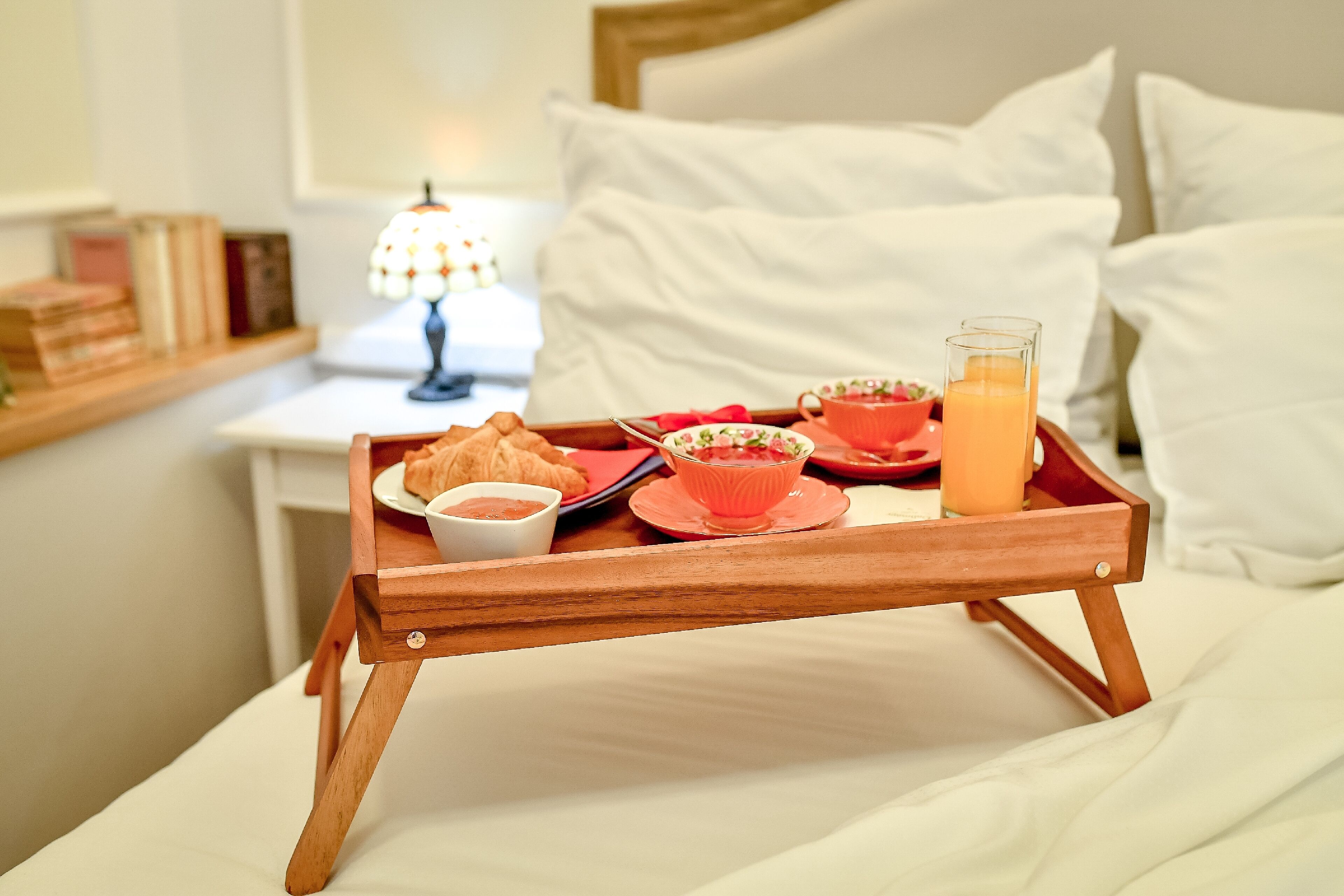 A breakfast tray with croissants, a bowl of fruit, a cup of tea, and a glass of orange juice on a bed with white bedding and pillows.