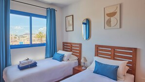 A hotel room with two beds, a large window with blue curtains, a wooden headboard, and a view of the city. - Casa Maria del Mar 2 Minutes From Supermarkets & 10 Minutes From Marina Rubicón (Playa Blanca)