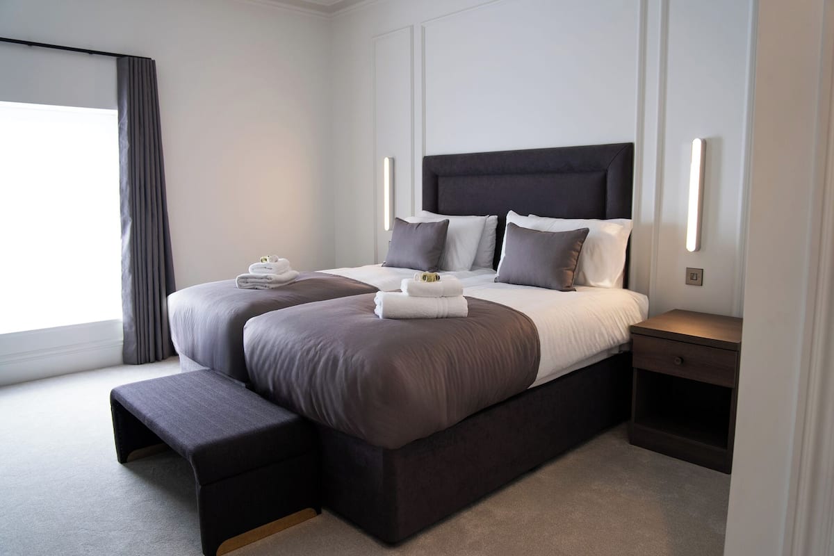 A modern hotel room with a large bed, a bench at the foot of the bed, and a bedside table with a lamp.