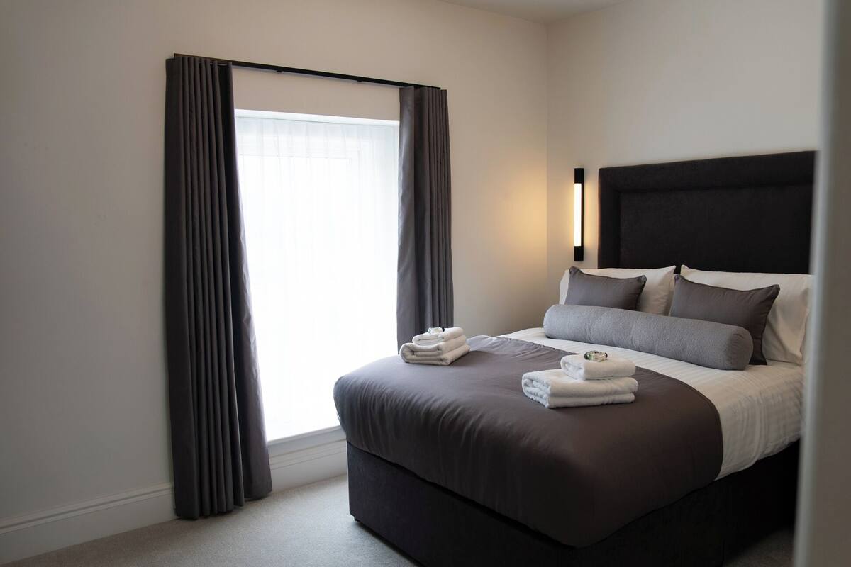 A modern hotel room with a large bed, dark curtains, and a headboard with built-in lighting.