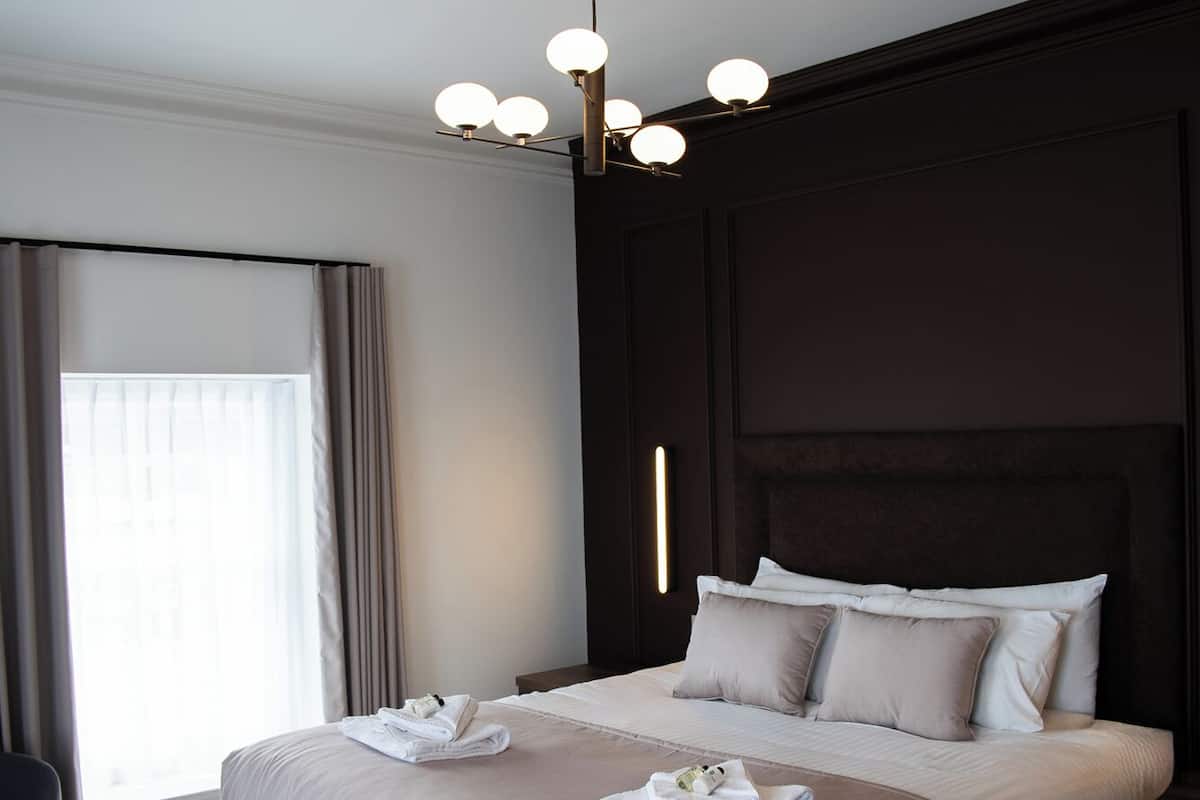 A modern hotel room with a large bed, a bench at the foot of the bed, a bedside table, and a hanging light fixture.