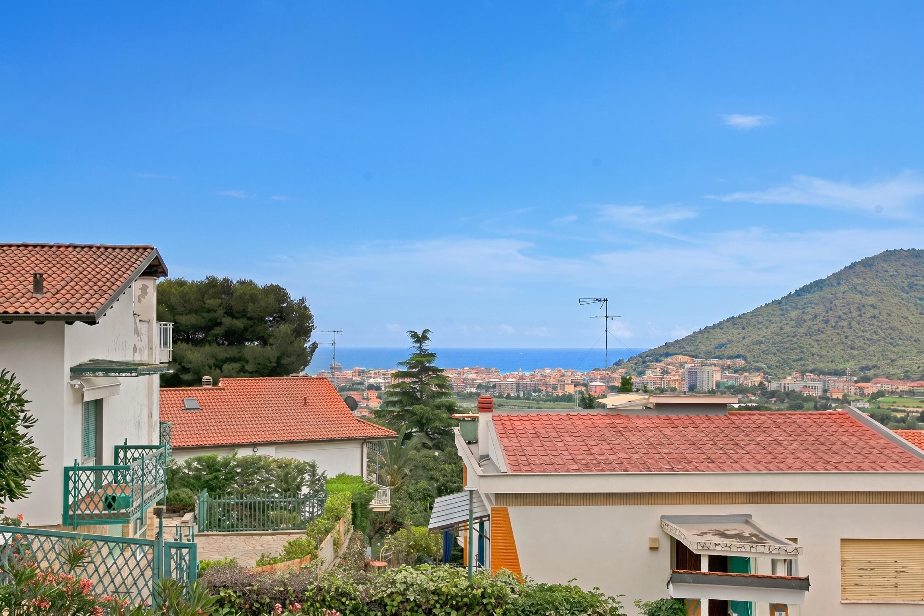 Ein Wohngebiet mit roten Ziegeldächern, Blick auf das Meer und eine Hügelkulisse im Hintergrund.