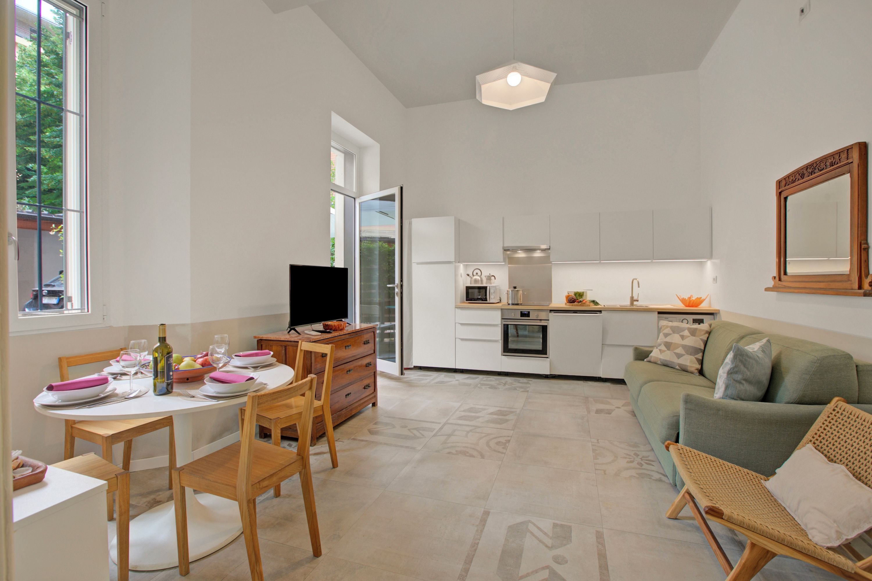 Uma cozinha moderna com armários brancos, uma sala de jantar com mesa e cadeiras redondas, e um sofá verde numa sala de estar ampla.
