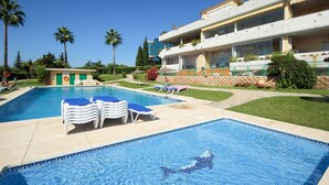 A swimming pool with lounge chairs, a building with balconies, and palm trees. - Penthouse @ Las Mimosas Cabopino (Marbella)