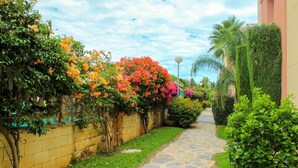 A stone pathway flanked by lush greenery and flowering plants, leading to a building with a yellow wall and a palm tree. - Penthouse @ Las Mimosas Cabopino (Marbella)