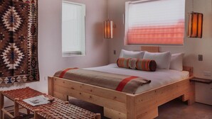 Een houten bed met een rood-beige deken, een geweven bankje en een wanddecoratie met een patroon.