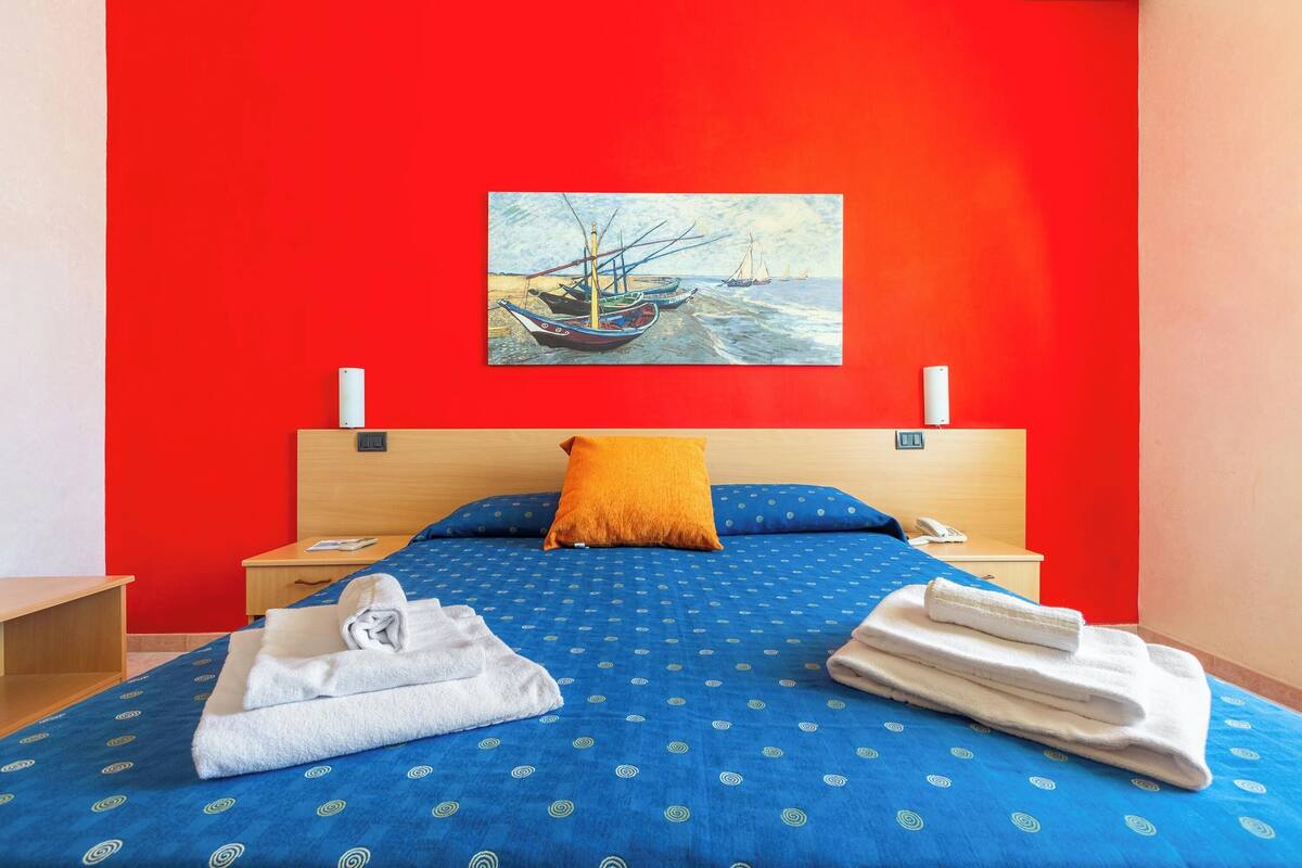 Een hotelkamer met een bed, een schilderij van boten aan de muur en een rode accentmuur.