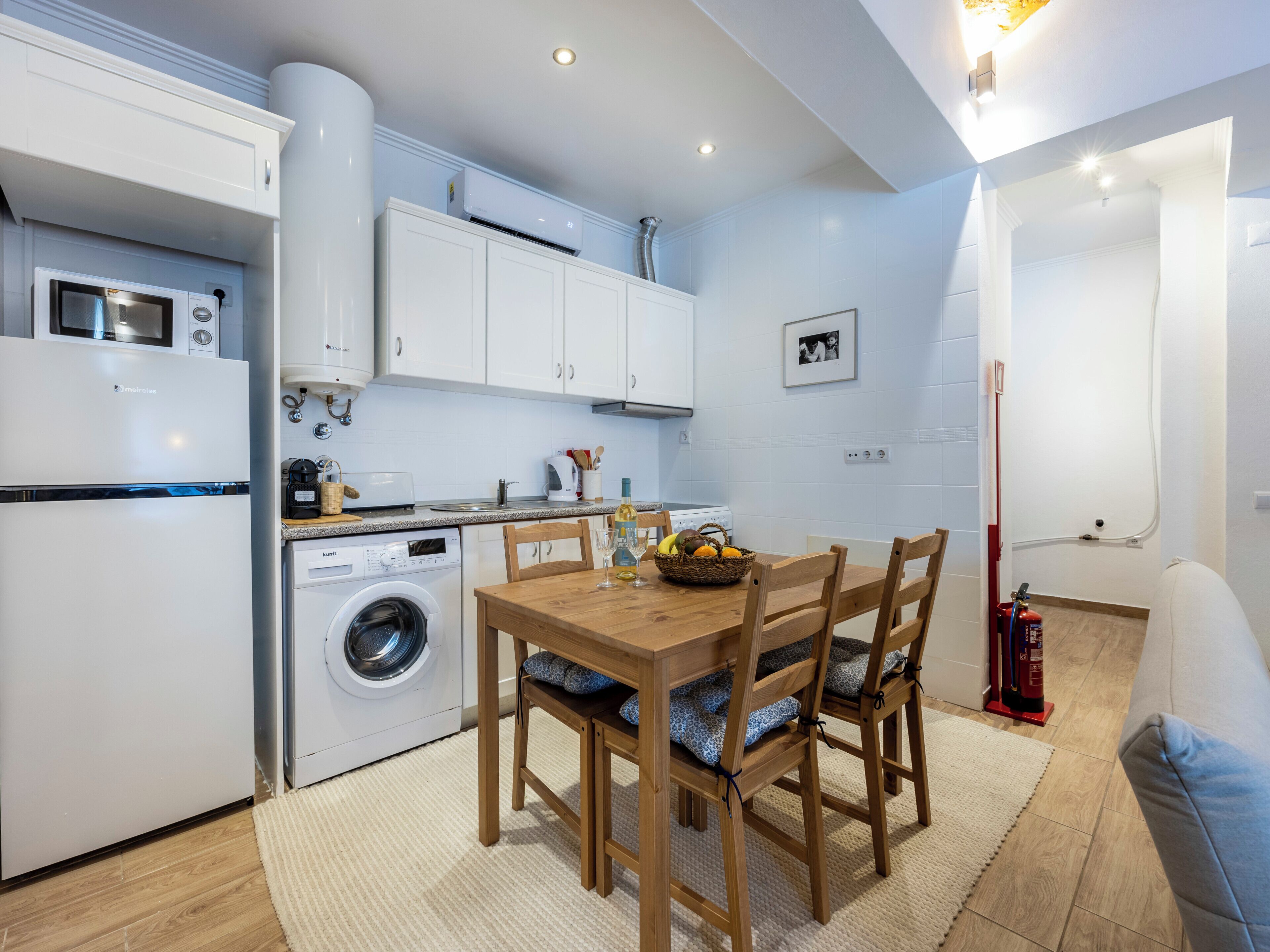 A compact kitchen with a washing machine, a microwave, and a stove. There is a wooden dining table with chairs and a fire extinguisher near the wall.