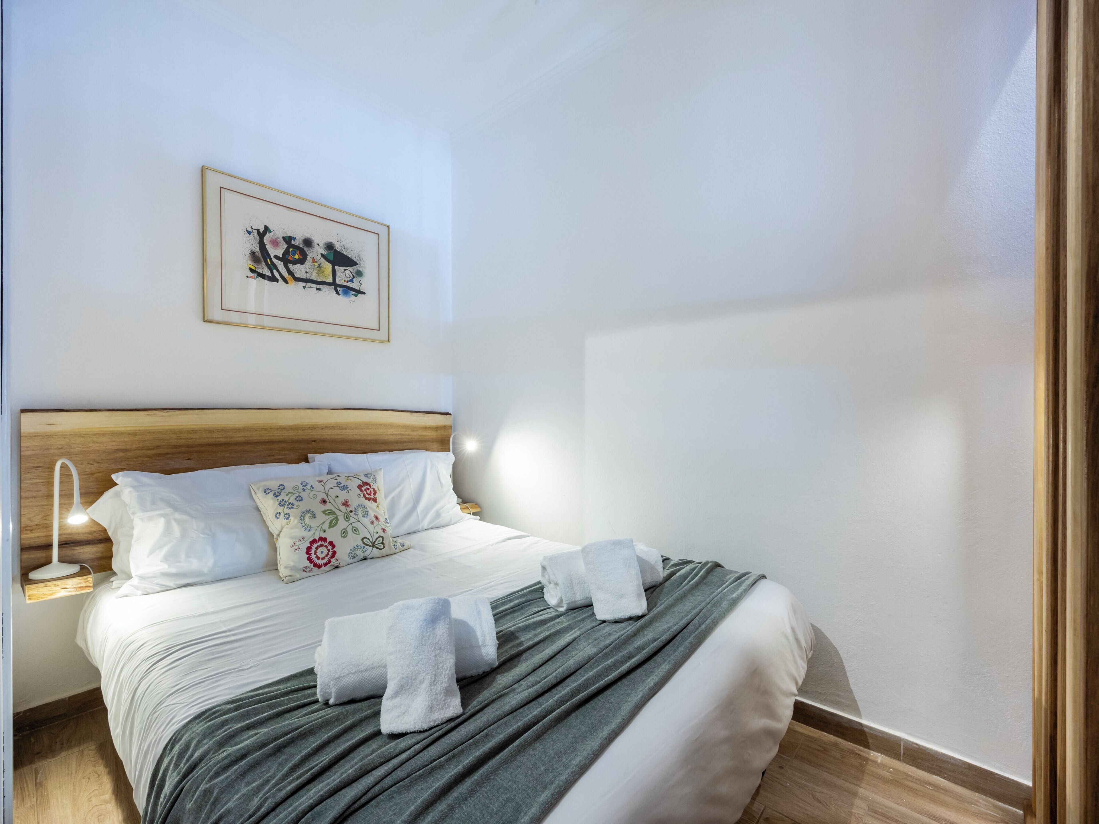 A neatly made bed with white linens, a green blanket, and two towels. There is a framed picture on the wall above the bed.