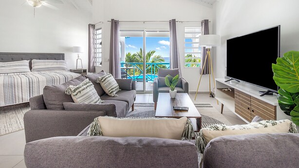 A modern hotel room with a bed, sofa, TV, and balcony with a view of the ocean.