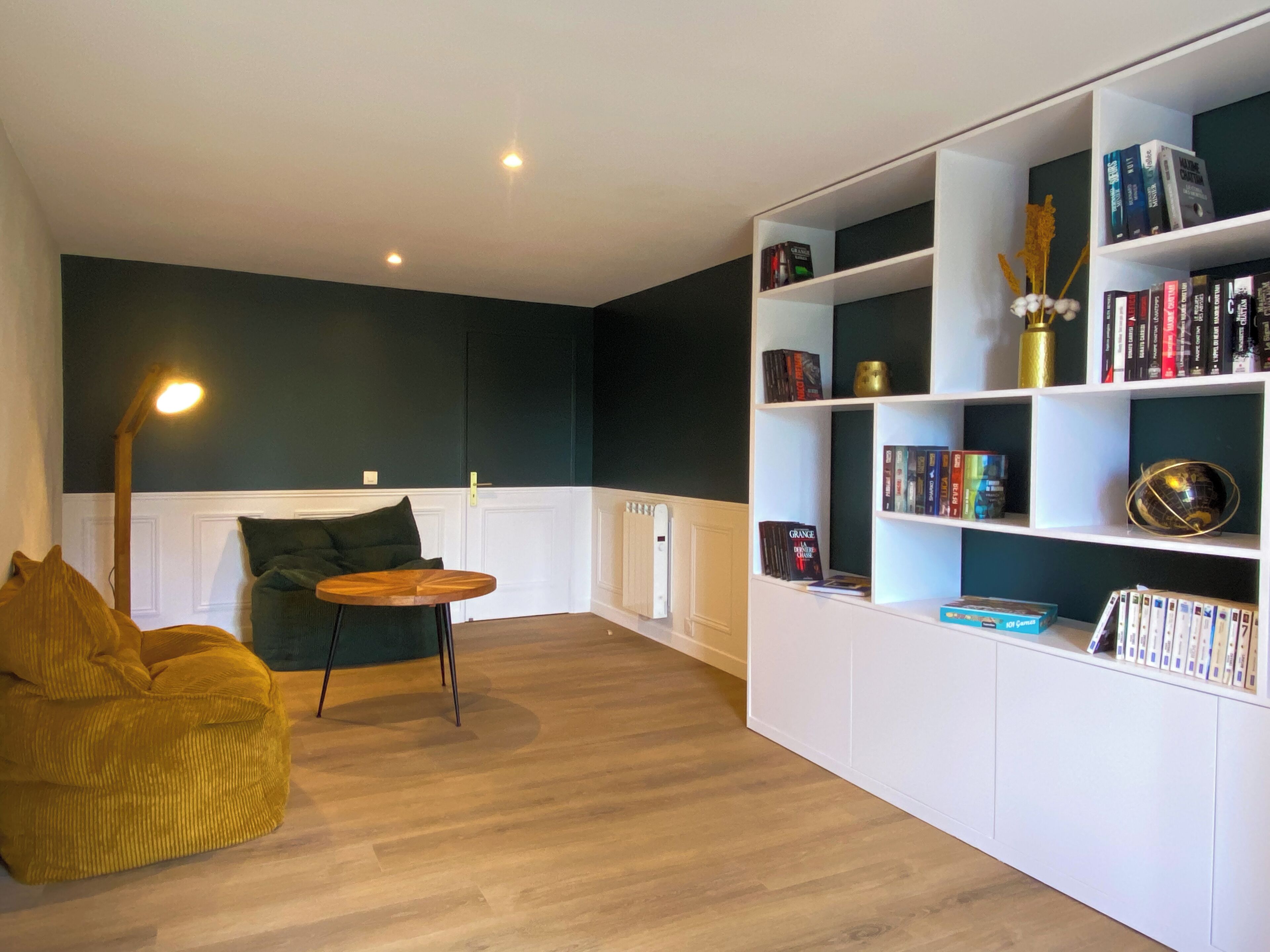 A modern living room with a dark accent wall, a wooden floor, a green sofa, a small round table, and a white bookshelf filled with books and decorative items.