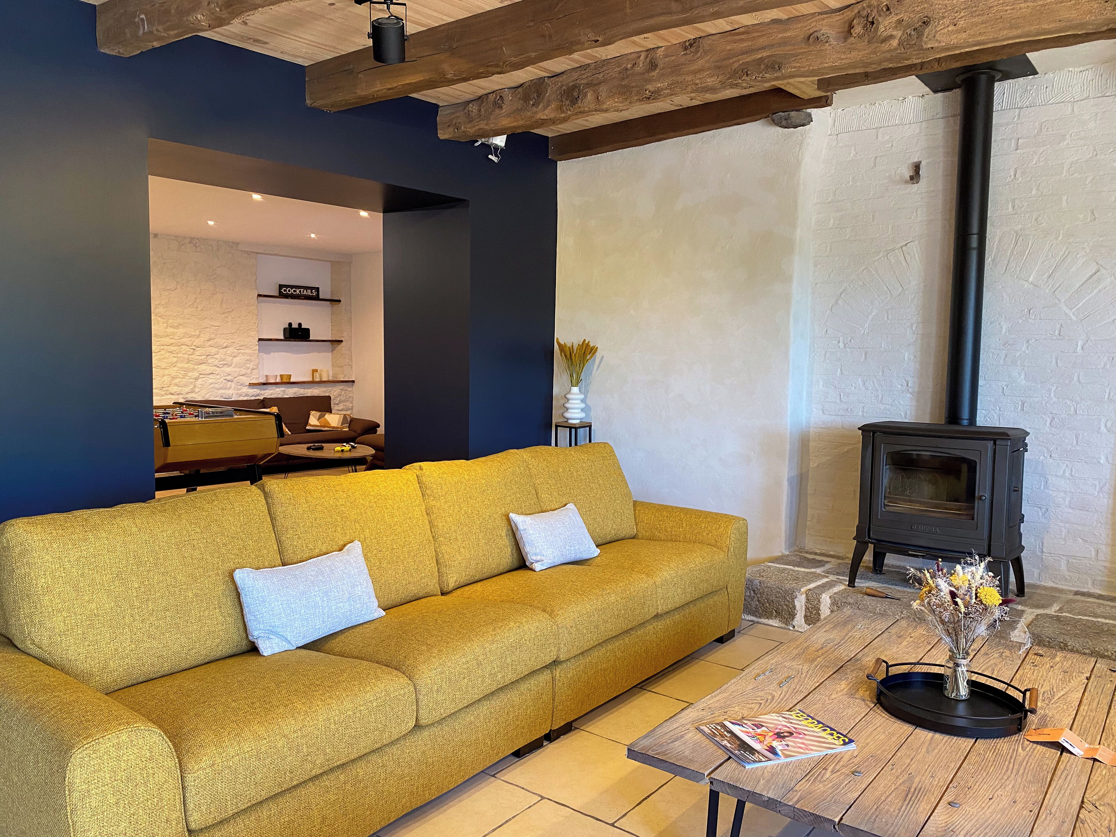 Una sala de estar con un sofá color mostaza, una mesa de centro de madera con revistas y un estufa de leña negra.